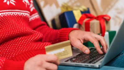 Pessoa fazendo compras online de Natal para aumentar o ticket médio, com presentes ao fundo e usando cartão de crédito.